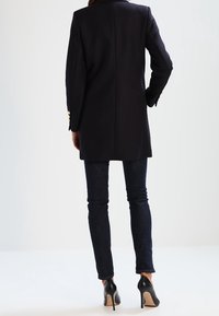 Marinblå lång kavaj i skräddarsydd stil med detaljer i guldknappar. Kombinerad med mörka slim fit jeans och svarta högklackade skor.