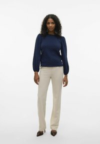 Pullover blu navy con texture e maniche a sbuffo, abbinato a pantaloni beige su misura e décolleté marroni a punta con borchie decorative.