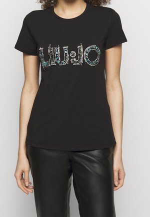 Kobieta nosząca czarną koszulkę z krótkim rękawem z logo LIU JO w niebieskich i białych kryształkach, w połączeniu z czarnymi skórzanymi spodniami.