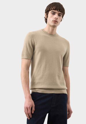 Giovane uomo che indossa un maglione beige a maniche corte e pantaloni scuri, in piedi davanti a uno sfondo chiaro uniforme.