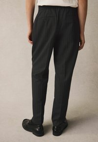 Pantaloni neri a righe con vita elastica e due tasche. Il modello indossa scarpe nere. La texture appare liscia e lucida.