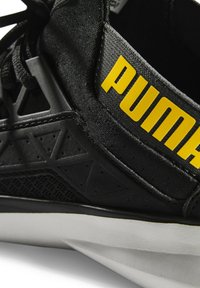 Zwarte sportieve schoen met gestructureerde stof, matte accenten en een rubberen zool. Heeft gedurfde gele 'PUMA' branding en geometrische ontwerpelementen.