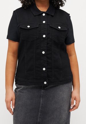 Person trägt eine schwarze kurze Jeansweste mit silbernen Knöpfen und zwei Brusttaschen, kombiniert mit einem dunkelgrauen Jeansrock.