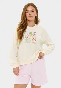 Krémový sweatshirt s farebnou grafickou potlačou nápisu "LA DOLCE VITA Italy." V kombinácii s svetloružovými šortkami.