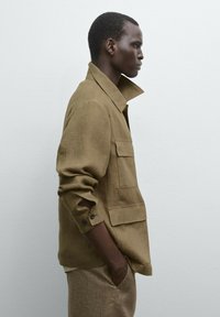 Brown linen jacket with a collar, two front pockets, and rolled sleeves. The fabric has a textured appearance, suitable for casual wear.