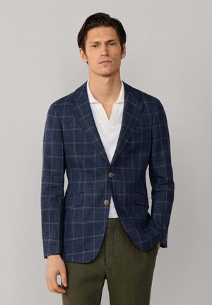 Man draagt een marineblauw geruit colbert, wit overhemd met open kraag en olijfgroene broek, staat met één hand in de zak.