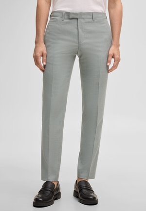 Uomo che indossa pantaloni eleganti su misura di colore grigio chiaro, mocassini neri in pelle e una camicia bianca infilata, in piedi davanti a uno sfondo semplice.