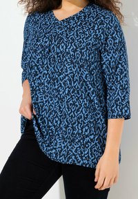 Marinblus med blå abstrakt mönster, V-ringning, trekvartsärm och en sidoklyvning. Kombinerad med svarta byxor.