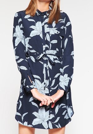 Femme portant une robe chemise bleu marine avec de grandes imprimés floraux blancs, boutonnée à l'avant, avec une poche poitrine et une ceinture nouée à la taille.