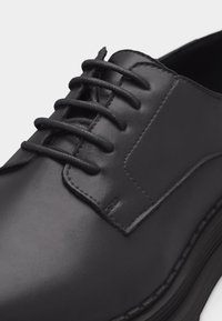 Μαύρο δερμάτινο επίσημο παπούτσι με κλειστή κορδόνια, ραμμένη λεπτομέρεια και παχύ πάτο, εμφανιζόμενο σε κοντινό πλάνο από την εμπρόσθια πλευρά.