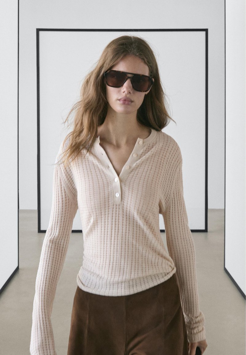 Femme portant des lunettes de soleil foncées, un haut beige à manches longues côtelé avec des boutons, et un pantalon marron, se tenant dans un espace de galerie minimaliste.