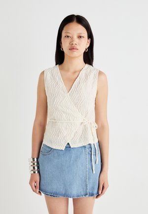 White textured wrap top with a V-neck, tie closure, and sleeveless design, paired with a light blue denim skirt.