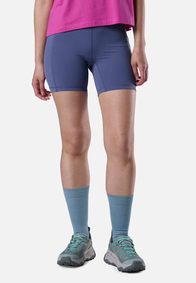 Persona che indossa pantaloncini sportivi blu, una maglietta rosa, calzini blu alti e scarpe da trekking grigio-verde, in piedi su uno sfondo bianco.