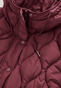 Veste rembourrée couleur prune avec texture matelassée, dotée d'un col montant, d'une capuche à cordon de serrage et de boutons-pression à l'avant.