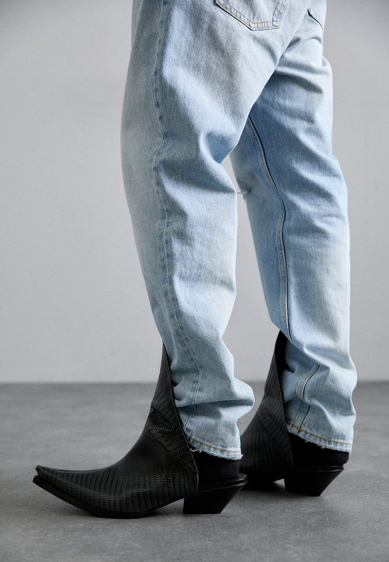 Bottes de cowboy noires avec un design à bout pointu et une surface texturée, associées à un jean droit bleu clair, le tout sur un fond gris.