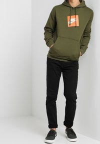 Olivgrön hoodie med en central orange Nike-logotyp, framficka, dragsko på huvan, kombinerad med svarta jeans och mörka sneakers.