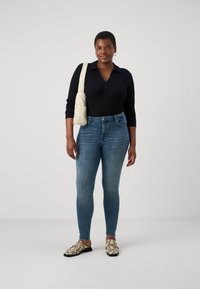 Black long-sleeve top, high-waisted blue skinny jeans, and beige patterned shoes. Accessories include a beige shoulder bag and hoop earrings.