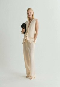 Chaleco beige entallado con escote en V profundo, combinado con pantalones de pierna ancha. La modelo sostiene un bolso negro texturizado, con un brillo sutil.