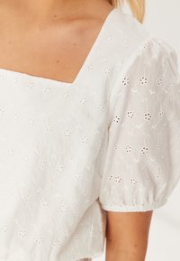 Top bianco con maniche a sbuffo e scollatura quadrata, caratterizzato da motivi floreali ricamati e piccoli intagli su un tessuto testurizzato.