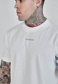 Bílý bavlněný tričko s kulatým výstřihem, krátkými rukávy a modrým logem "SIKSILK" na prsou. Hladký materiál, volný střih.