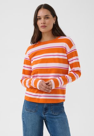 Femme aux longs cheveux bruns porte un t-shirt à manches longues rayé horizontalement orange, rose et blanc et un jean bleu, debout, les mains jointes.