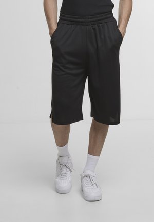 Mężczyzna w czarnych spodenkach do kolan, białych skarpetkach i białych sportowych butach na sznurowadła, stojący z rękami w kieszeniach na białym tle.