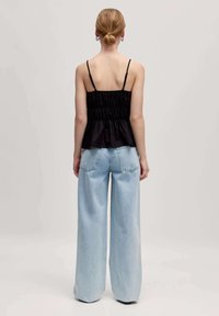 Zwarte peplumtop met spaghettibandjes en geplooide textuur, gecombineerd met wijde lichtblauwe denimjeans. Achteraanzicht getoond.