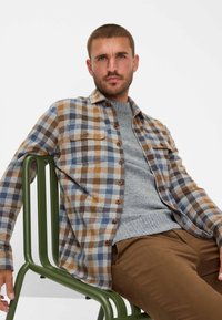 Geruite shirt in beige, blauwe en bruine tinten; met knopen, gemaakt van stof met een zachte textuur; gedragen over een lichtgrijze trui.