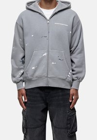 Felpa grigia con zip e tasca frontale, decorata con dettagli di schizzi di vernice blu, rosa e bianca. Include un cappuccio con cordino e polsini a coste.