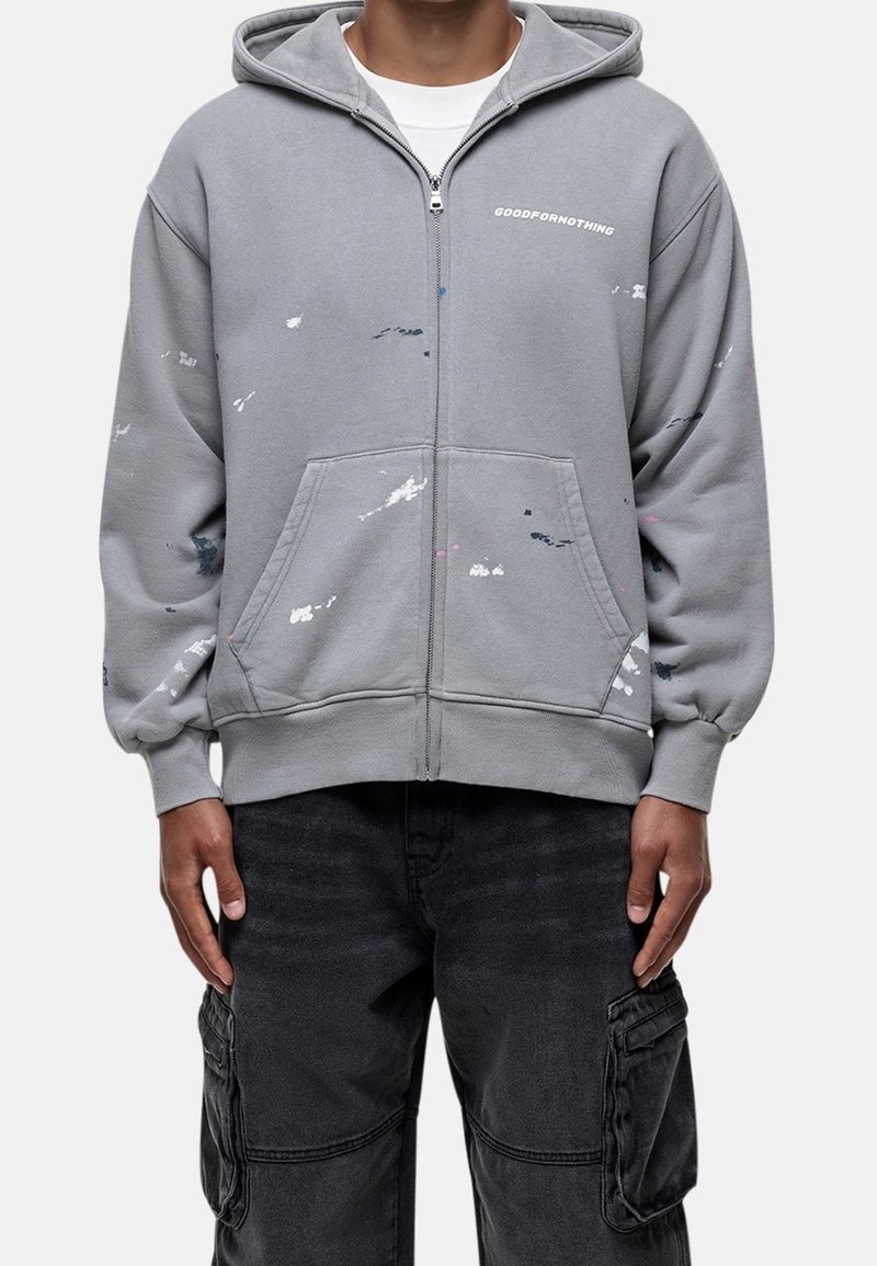 Felpa grigia con zip e tasca frontale, decorata con dettagli di schizzi di vernice blu, rosa e bianca. Include un cappuccio con cordino e polsini a coste.