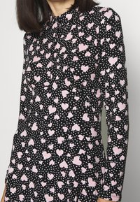 Svart tygklänning med ett mönster av rosa hjärtan och små vita hjärtan över hela. Långa ärmar och en knäppt krage.