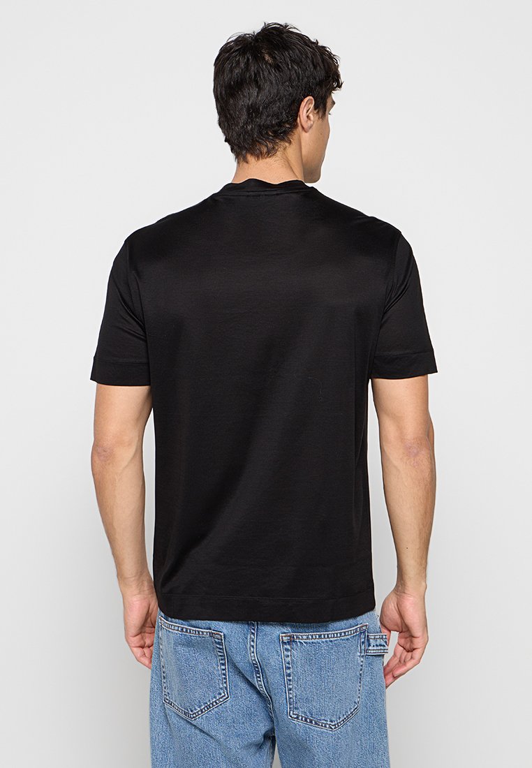 Hombre con camiseta negra lisa de manga corta y vaqueros azules, de espaldas frente a un fondo liso y claro.