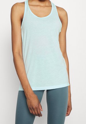 Kvinde iført en lyseblå, ærmeløs tanktop og mørkeblå leggings, stående foran en ensfarvet hvid baggrund.