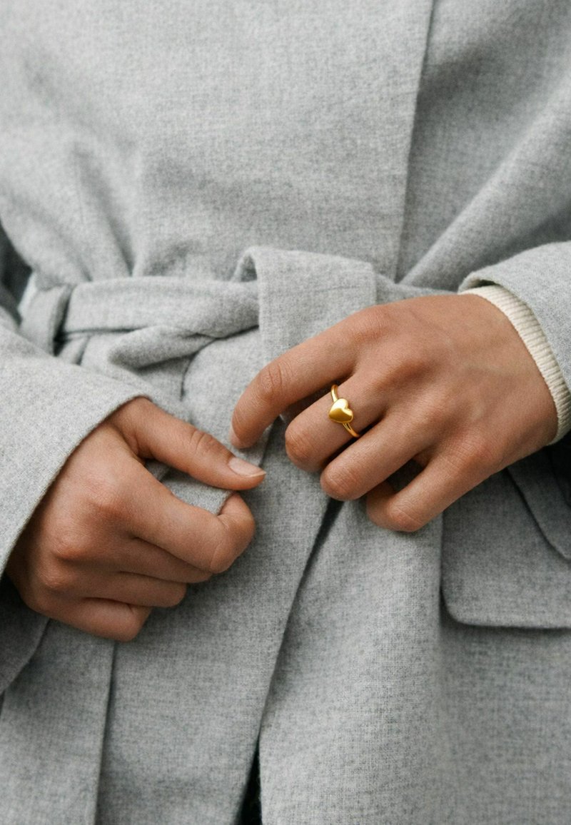 Des mains attachant la ceinture d'un manteau en laine gris clair, avec une bague en forme de cœur en or à l'annulaire de la main droite.