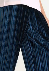 Marineblå plisserede fløjl bukser med en blød glans, der har skarpe lodrette linjer og en afslappet pasform. Nærbillede fremhæver tekstur og farver.
