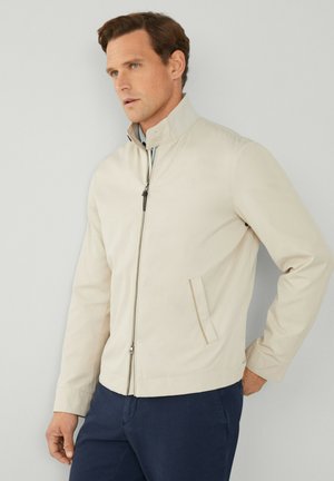 Veste légère beige avec col montant, fermeture éclair à l'avant, poches latérales et texture lisse. Portée par-dessus une chemise bleu clair.
