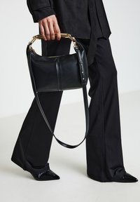 Schwarze Lederhandtasche mit goldfarbenem Schlaufenhenkel und verstellbarem Riemen, mit glatter Textur und Reißverschluss.