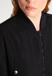 Czarna kurtka bombka z zamkiem z przodu, ściągaczem przy kołnierzu oraz kieszenią z metalowym zatrzaskiem. Gładka faktura i lekki materiał.