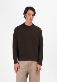 Brauner Strickpullover mit geripptem Halsausschnitt und Bündchen, im lässigen Schnitt. Getragen mit hellbeigen, strukturierten Hosen.