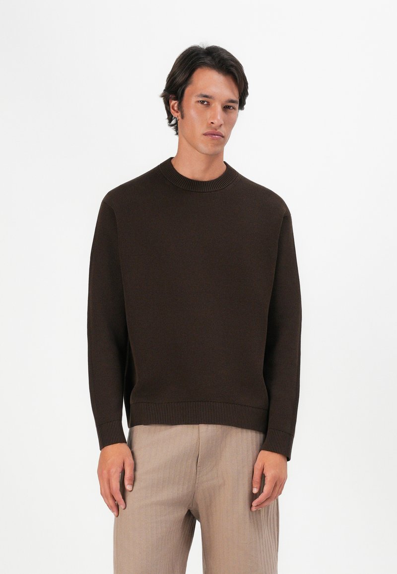 Brauner Strickpullover mit geripptem Halsausschnitt und Bündchen, im lässigen Schnitt. Getragen mit hellbeigen, strukturierten Hosen.
