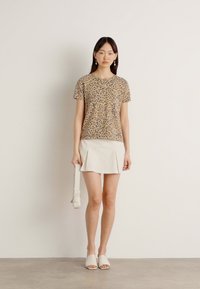 Kortermet skjorte med leopardmønster i beige, kombinert med et hvitt plisserte skjørt. Modellen bærer en hvit crossover-veske og slip-on sko.