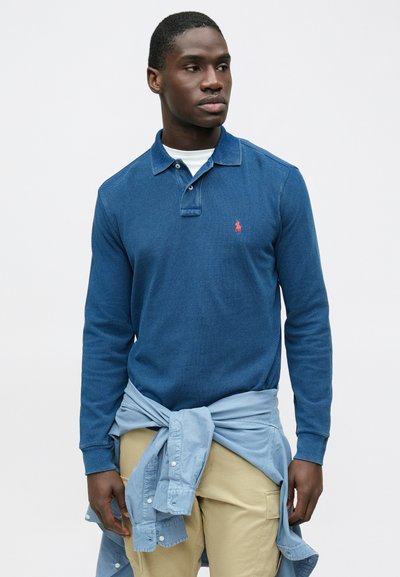 Temno modra polo majica z dolgimi rokavi in ovratnikom, z majhnim rdečim logotipom na prsih; svetlo modra majica vezana okoli pasu.