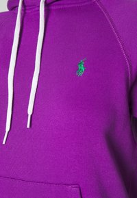 Lila hoodie i mjuk bomull, med vita dragsnören, känguruficka och en liten grön polo-logotyp på bröstet.