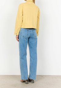 Donna che indossa jeans azzurri chiari, un giubbotto giallo corto e sandali con tacco a motivo serpente, in piedi rivolta di spalle contro un muro bianco.