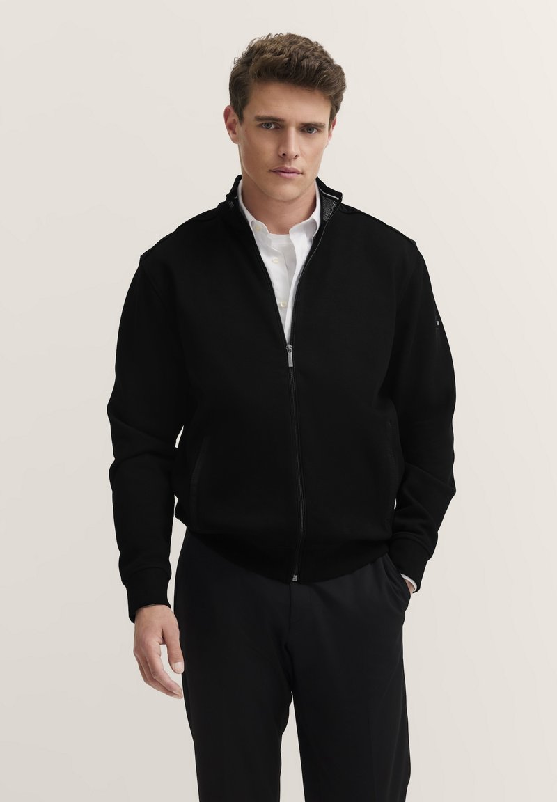 Schwarze Zip-Jacke mit hohem Kragen, aus weichem Stoff gefertigt. Verfügt über seitliche Taschen und eine glatte Textur. Getragen über einem weißen Hemd.