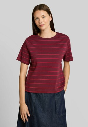 Jeune femme aux cheveux bruns raides porte un t-shirt manches courtes bordeaux à fines rayures rouges et une jupe bleu foncé avec des poches devant.