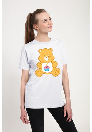 Ung kvinde iført hvid T-shirt med et gult teddybjørn, der holder en cupcake med et tændt lys trykt på, ser op og smiler.