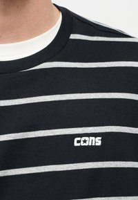 Κοντινό πλάνο ενός ατόμου που φοράει μαύρο και γκρι ριγέ T-shirt με λευκό γιακά και λευκό λογότυπο "cons" με αστέρι.