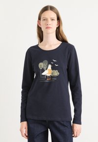 Tume sinise värvi pikkade käistega T-särk, mis on valmistatud puuvillast, sisaldades linnu graafikat oranžide ja roheliste detailidega ning taustal puud.