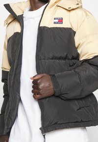 Svart och beige pufferjacka med huva, med dragkedja framtill, elastiska ärmslut och ett litet logopatch på bröstet.